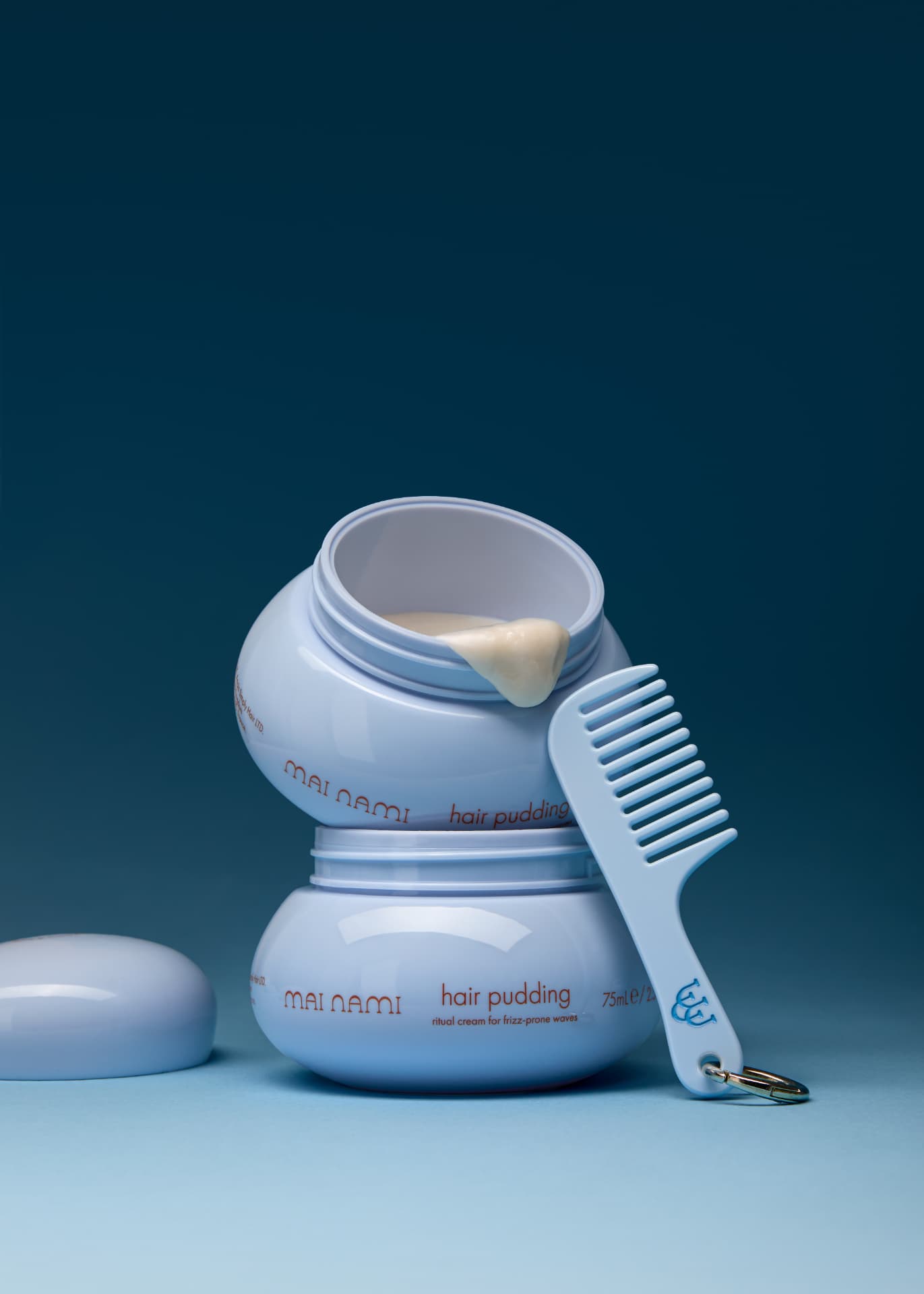 Stacked Mai Nami Hair Pudding pots with a wide-tooth comb on a deep blue backdrop